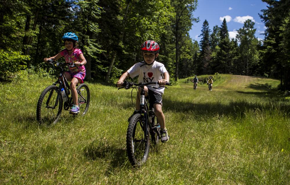 two children and adults ride bikes through a lush field surrounded by forest.