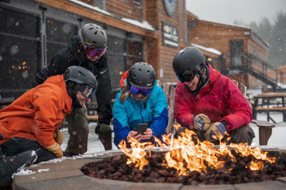 a group of skiers watches a video on their phone at an outdoor picnic table.