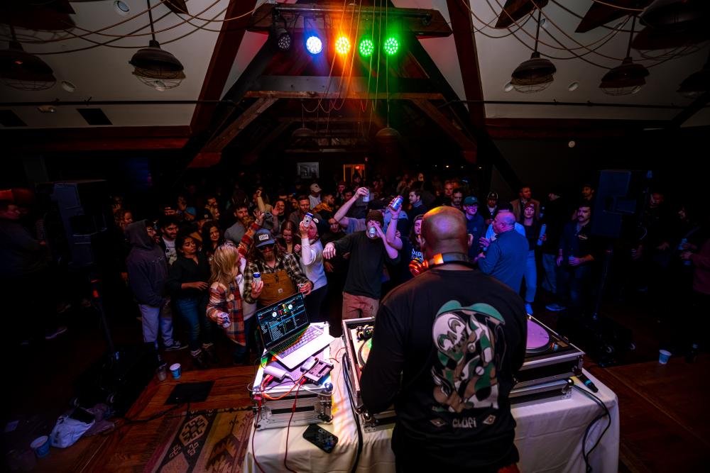 A DJ performs in front of a small crowd in a rustic indoor room.