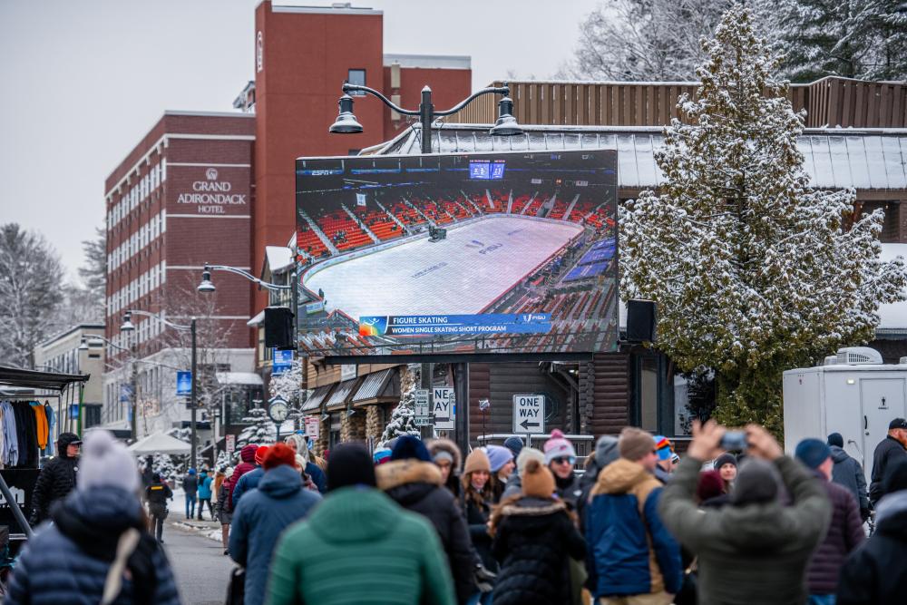 Pedestrians fill a street in front of a brick hotel and jumbotron showing a hockey game.