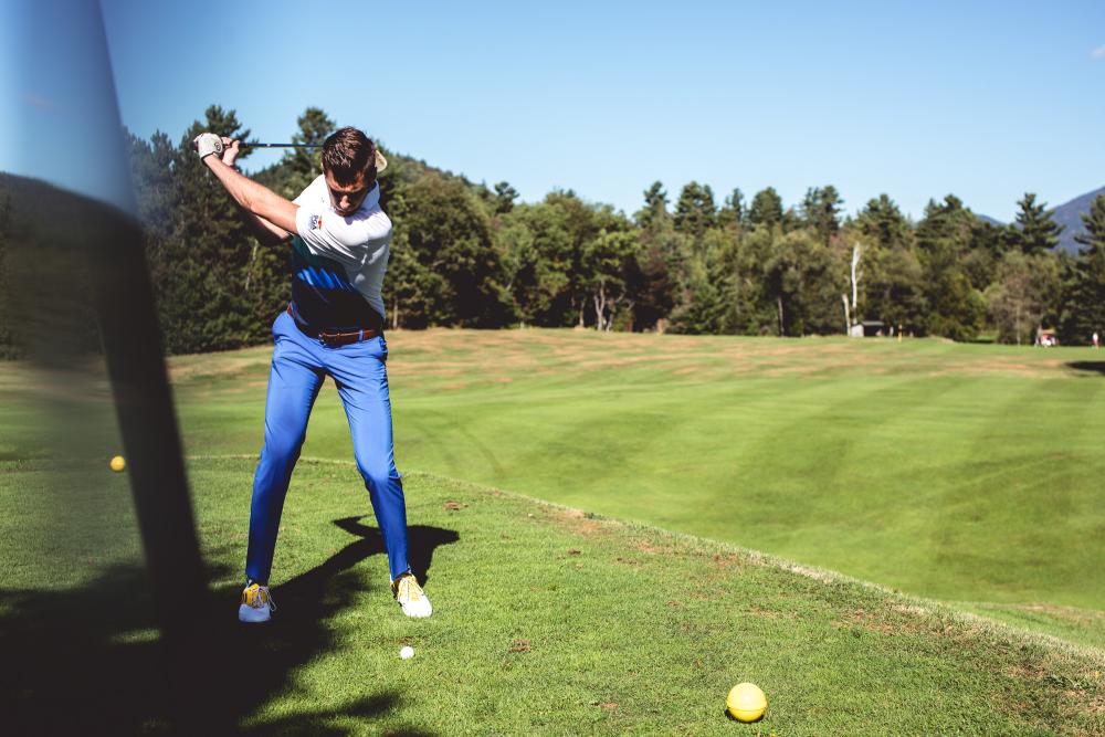 A man raises his club to hit a golf ball in front of a tree-line.