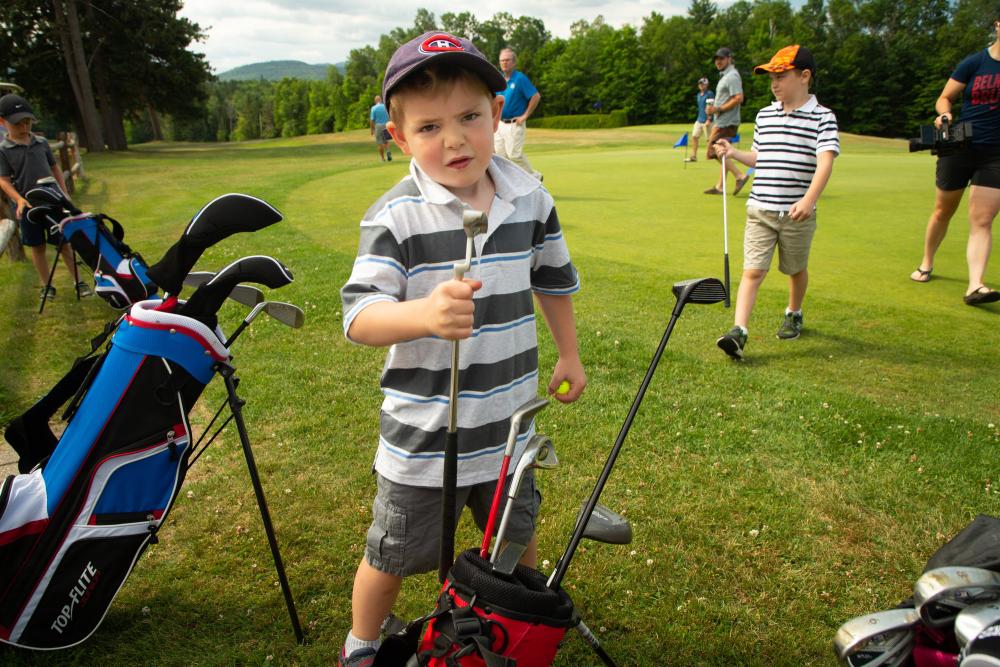 a young boy in a striped shirt holds his clubs at the golf course with other golfers around,