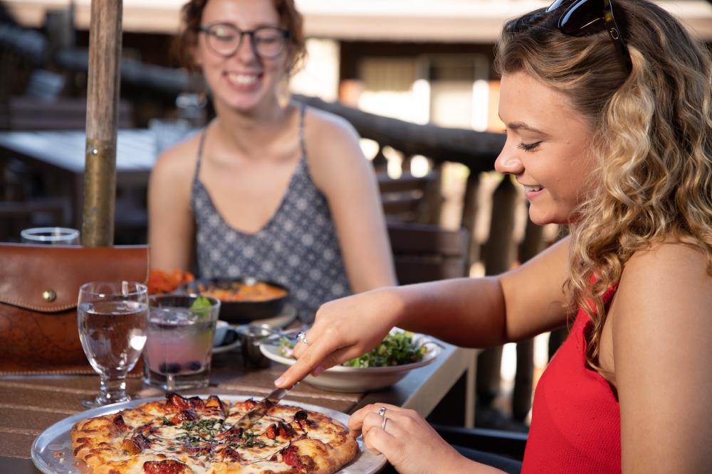 A woman dines with a friend in summer while cutting into a pizza.