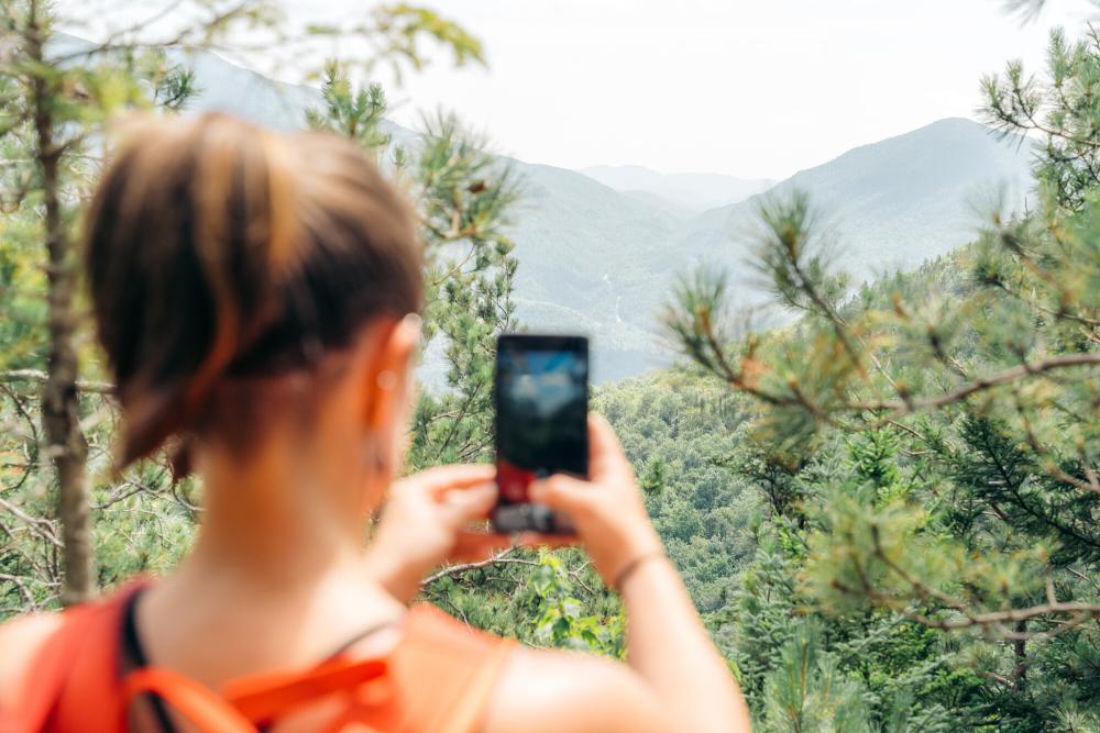 A woman in a tanktop takes a picture of a nearby High Peak in the woods.