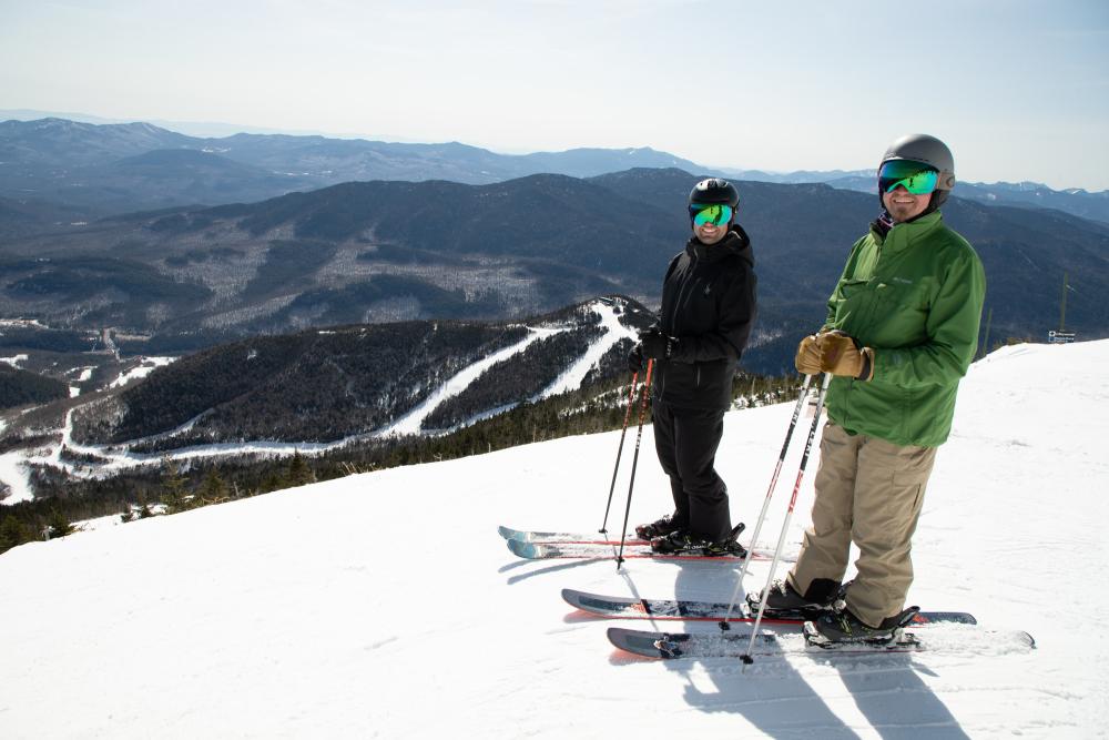 Two skiers pose on a snowy peak high above a mountain valley.