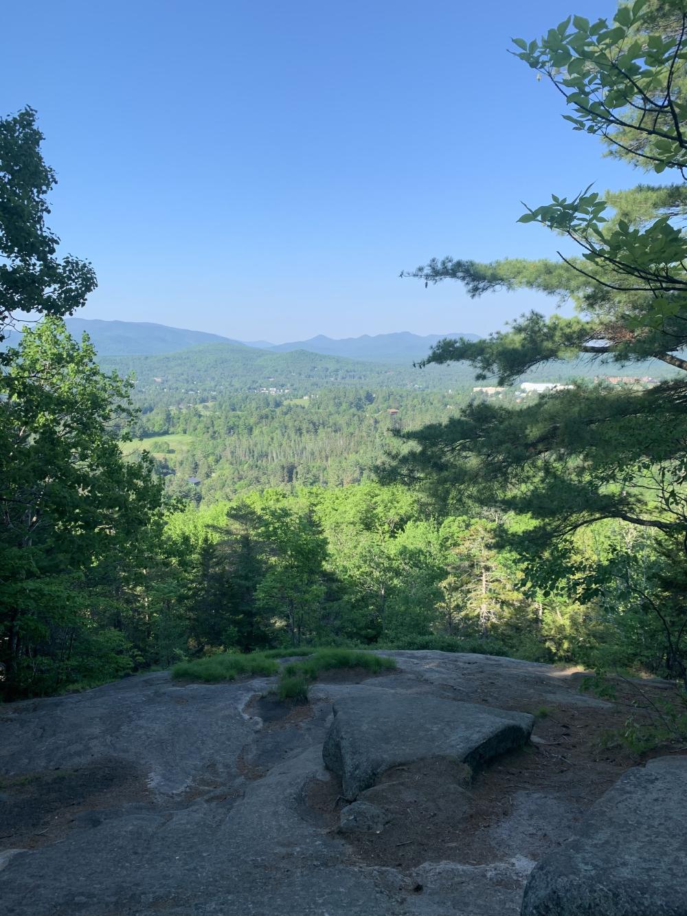 A rocky ledge offers a view of mountains and forest.