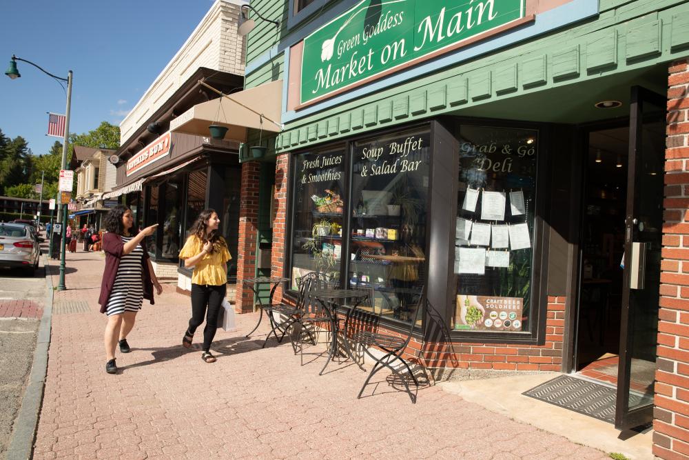 Two women walk down the brick sidewalk on Main Street near the Green Goddess Market on Main storefront with a green sign with white letters.