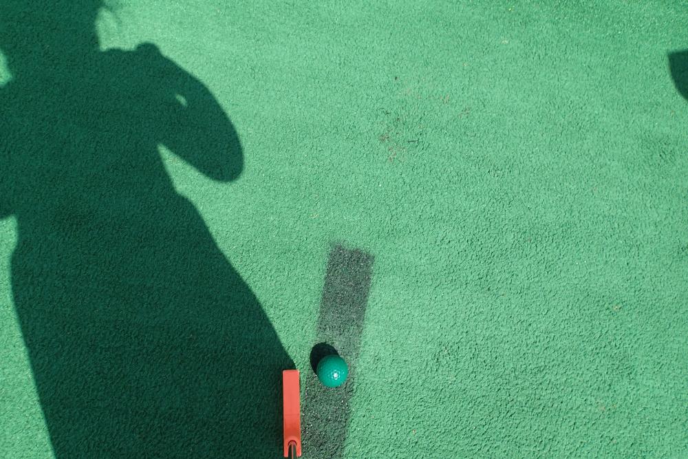 Self-portrait at Boots and Birdies. Photo by Evie, age 6. An image of artificial turf on a mini golf course, with a putter, golf ball, and the shadow of a little girl.