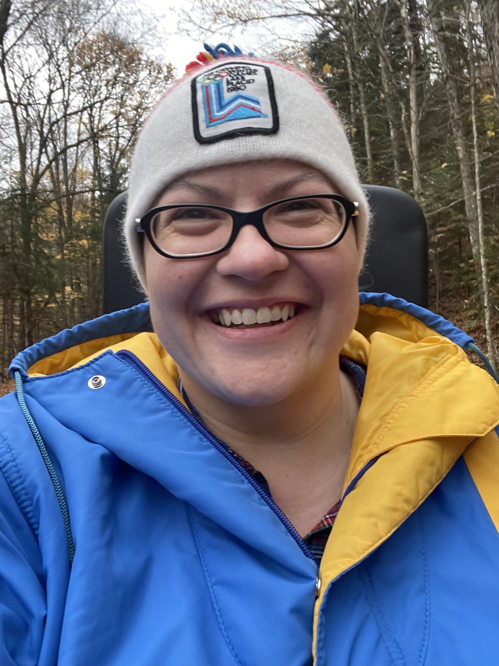 A woman in a bright blue and yellow parka and 1980 Lake Placid Olympic winter hat grins on a ride.