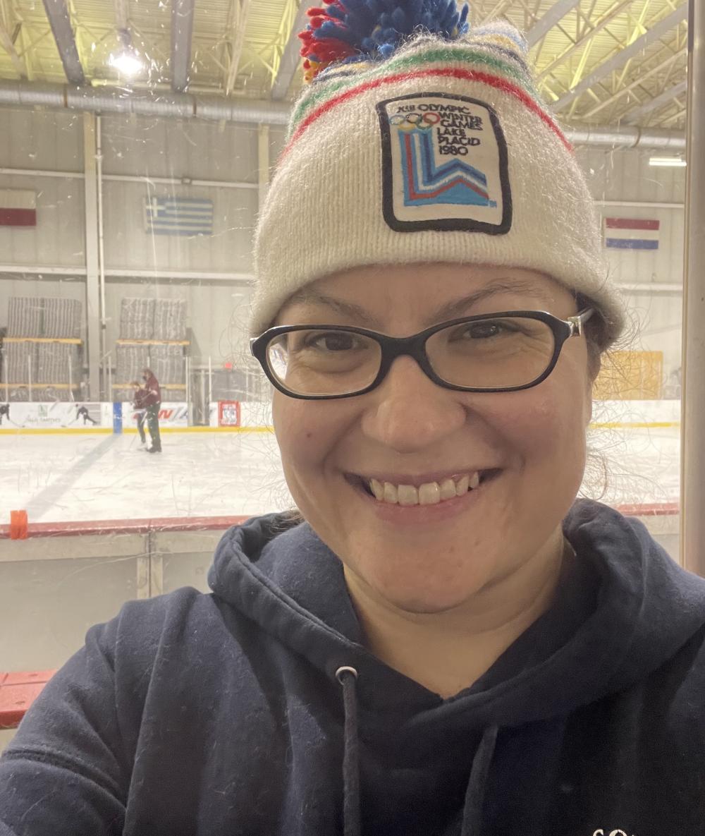 A woman in a 1980 Lake Placid Olympic winter hat smiles in front of an indoor skating rink.