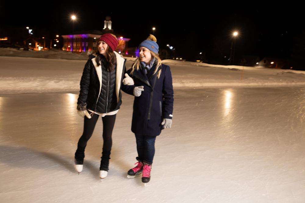 A pair of women in ice skates laughing on an outdoor rink in the evening.