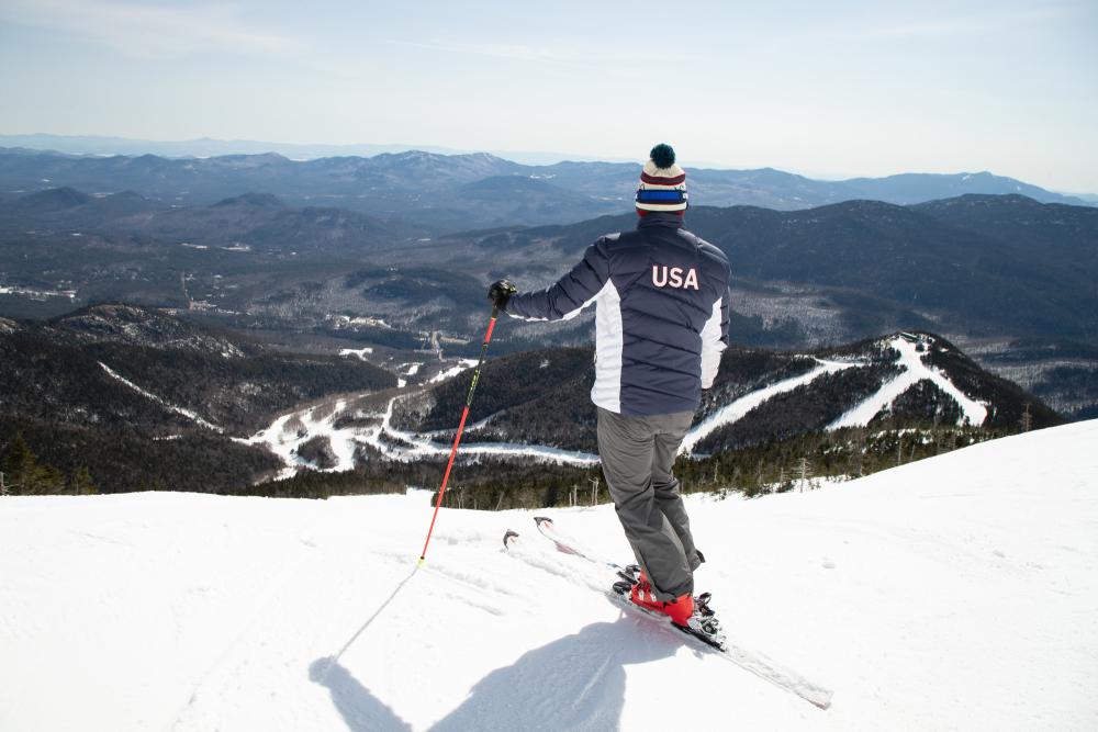 A skier in a red, white, and blue USA ski jacket poses on the edge of a run on a snowy mountain peak.