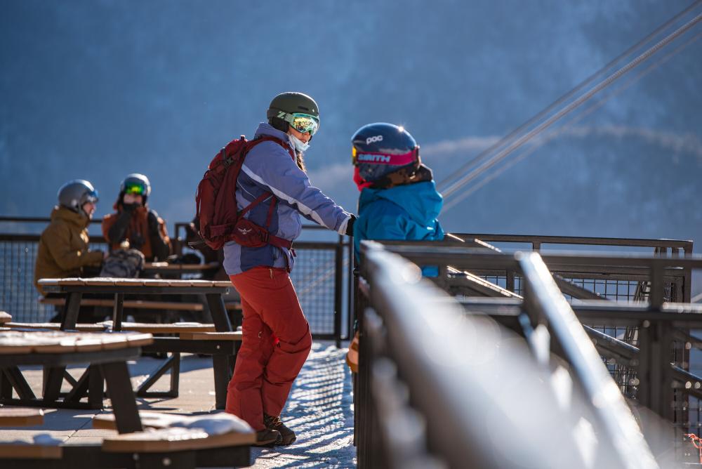 Two people talk outdoors on a patio dressed in ski gear, with helmets and jackets and snowpants.