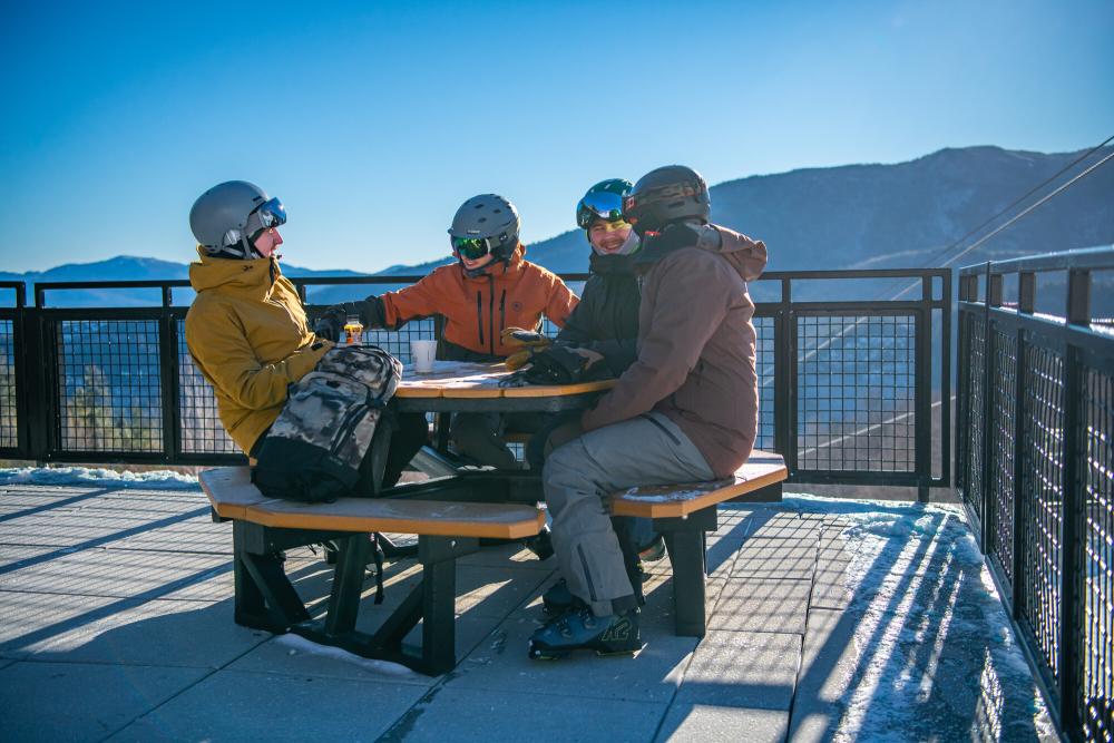 Four friends smile and laugh at an outdoor picnic table on a patio while dressed in ski clothes.