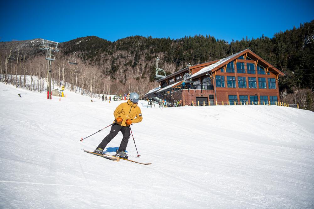 A skier in a yellow jacket goes down the mountain with a new Legacy Lodge in the background.