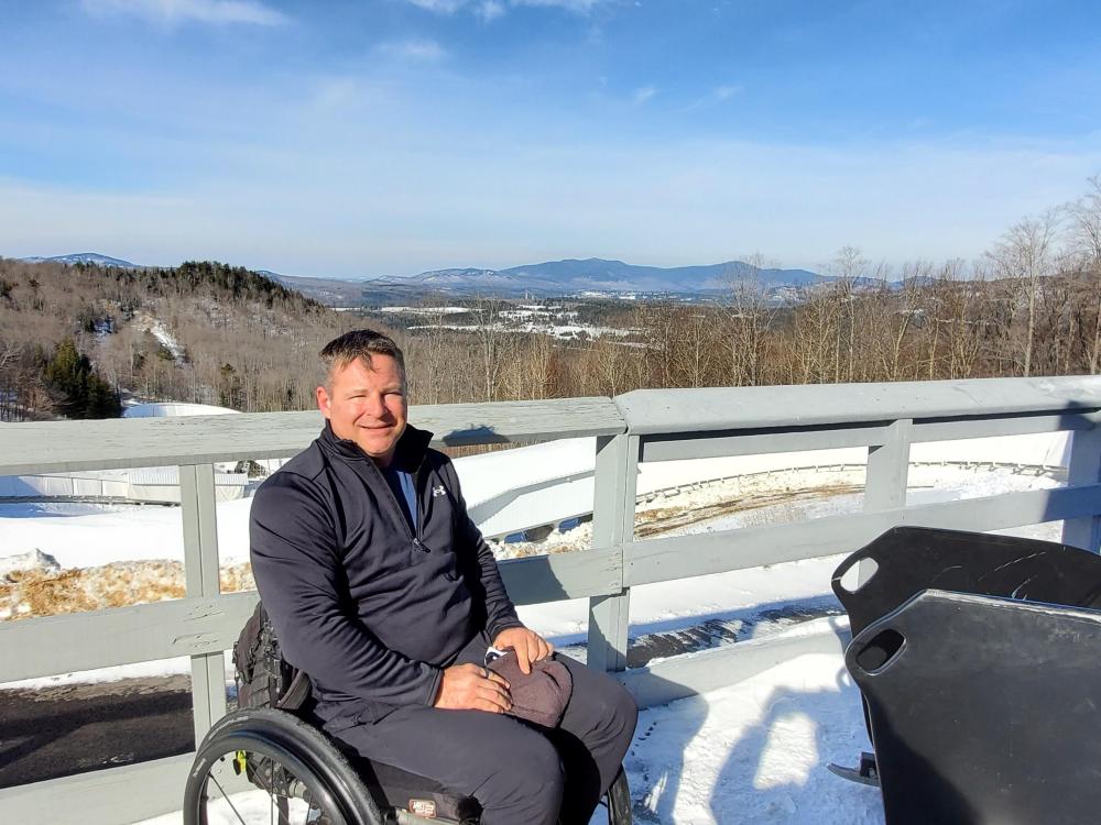 Adaptive athlete, David Christopher, visiting Lake Placid for a day of training on the bobsled track. Image courtesy of David Christopher. David Christopher in his wheelchair smiling with a wintery Lake Placid behind him. Image courtesy of David Christopher.