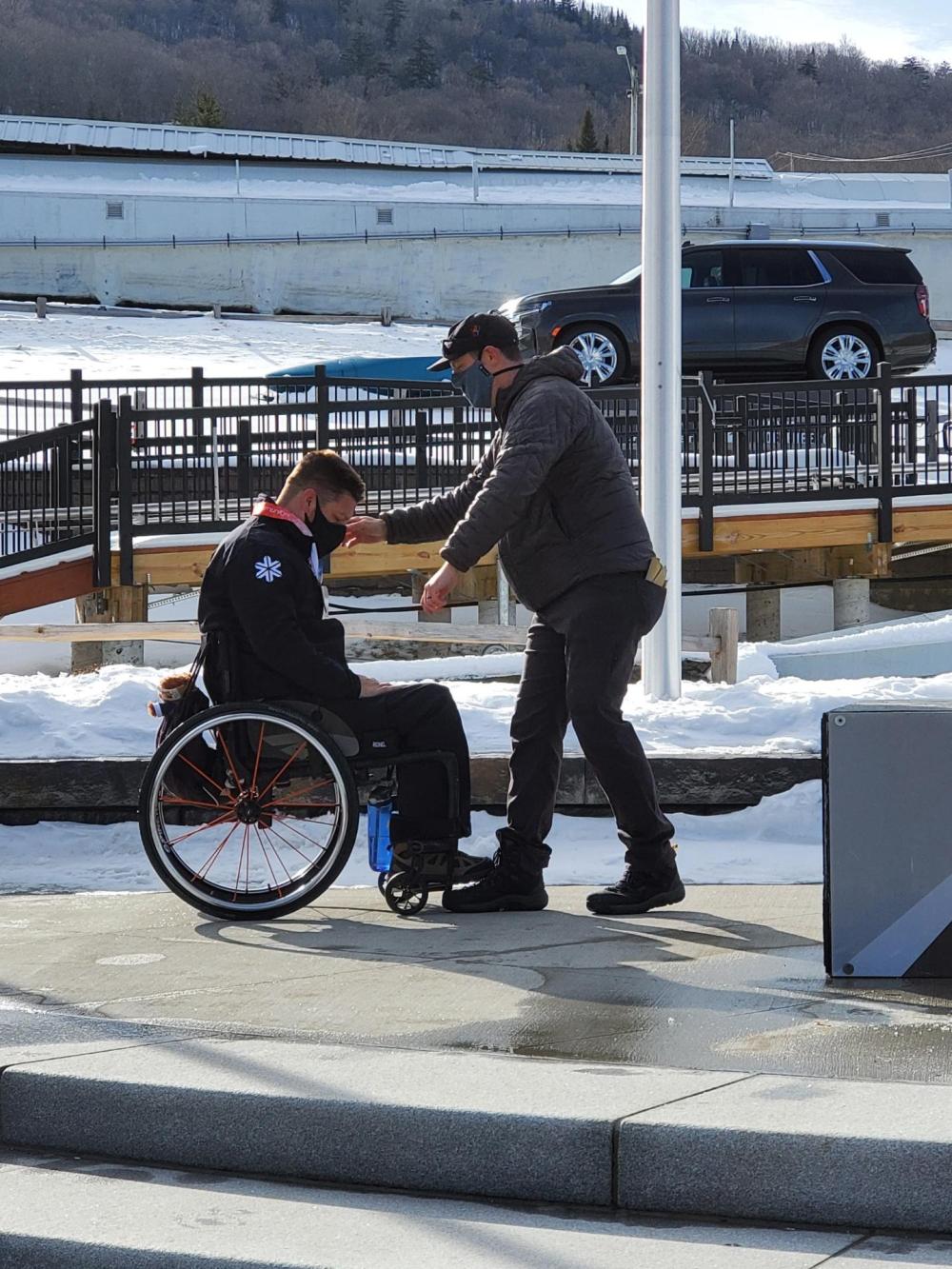 David Chrstopher arrives at the bobsled track in Lake Placid for a day of training. Image courtesy of David Christopher. A man stands beside a man in a wheelchair to help him along a sidewalk. Image courtesy of David Christopher.