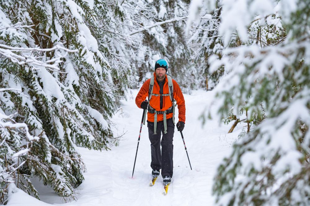 A man with a beard and a red jacket skis on a narrow trail lined with snow covered pine trees.