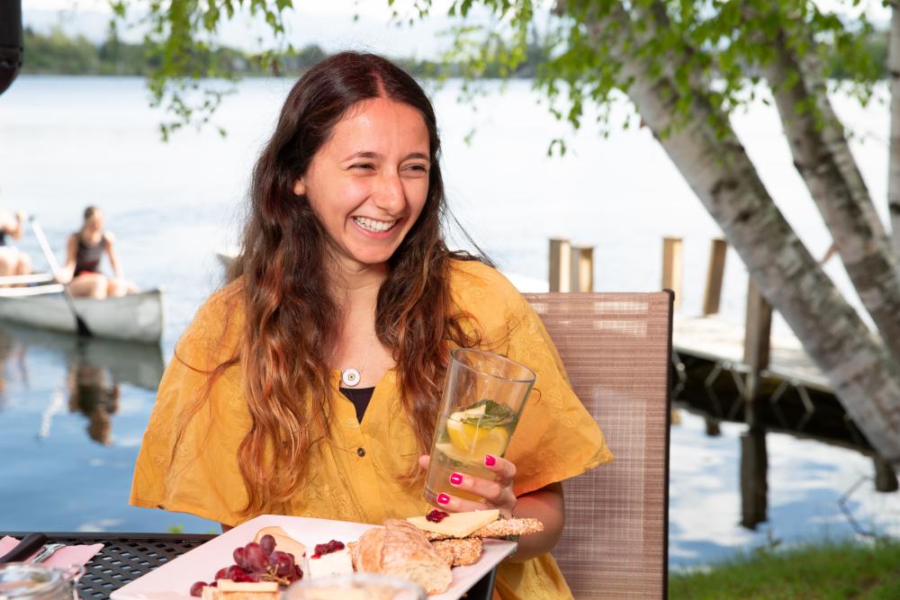 A young woman smiles widely at a table of food, while holding a drink in front of a lake with canoeists.