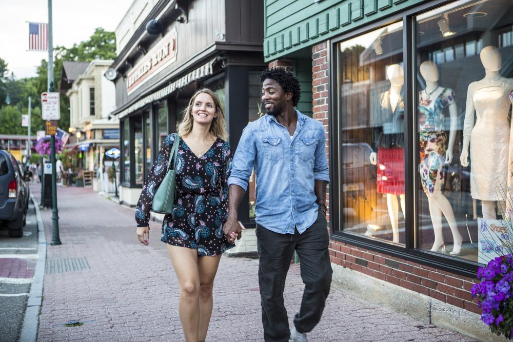 A man and a woman walk down a shop-lined sidewalk.