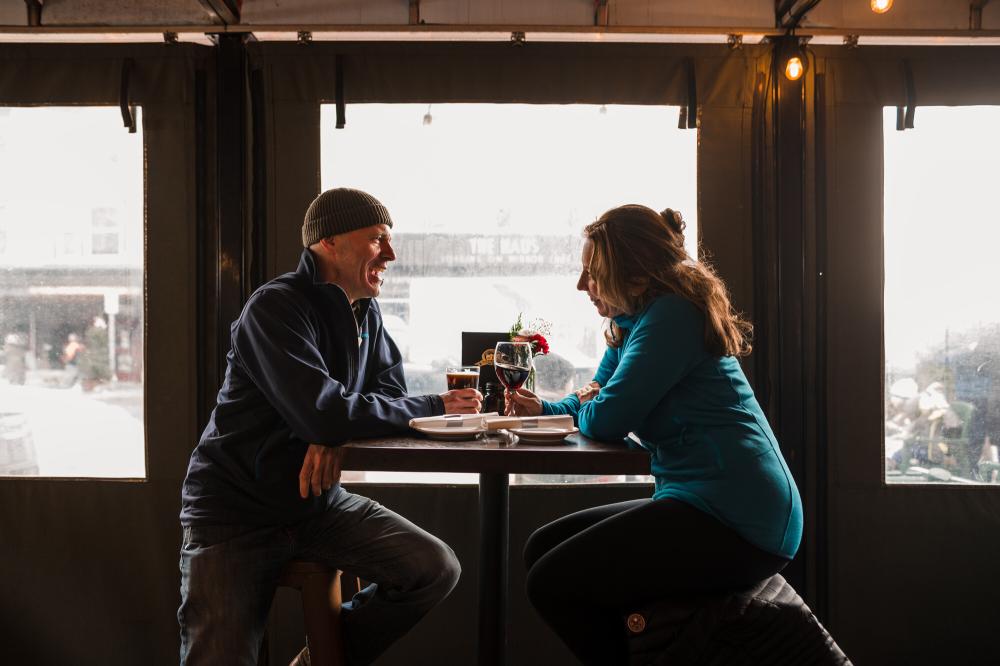 A couple sits on a covered porch and enjoy drinks while it rains outside.