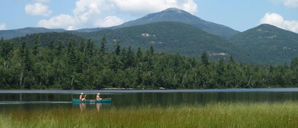 A green canoe with two people paddles on a calm lake with a large mountain in the background.