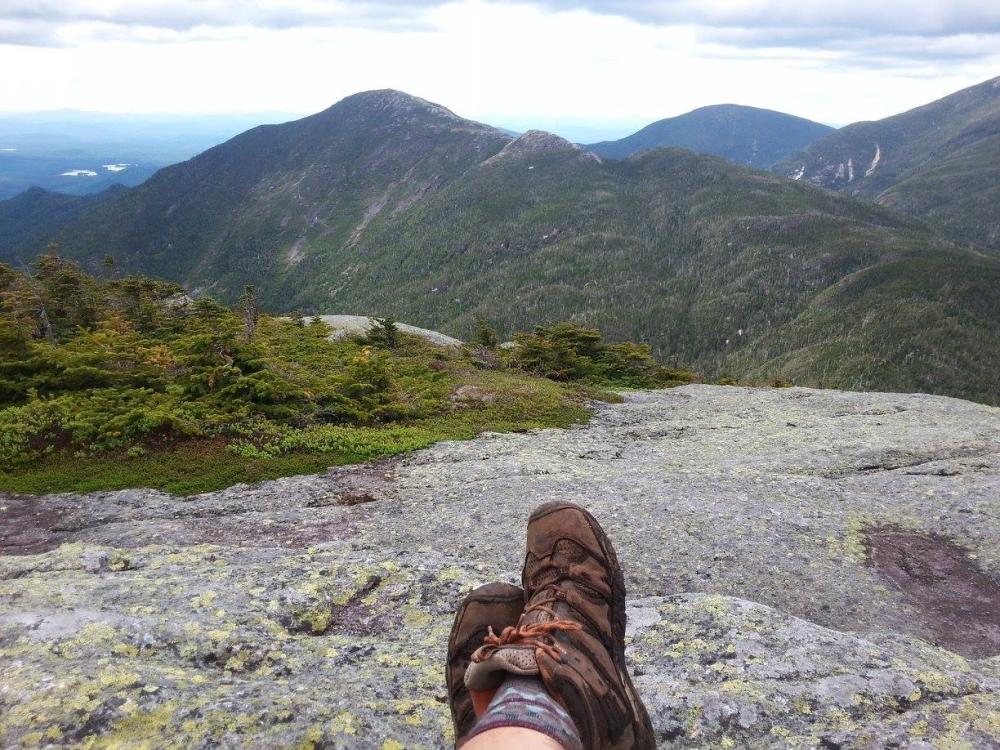 A hiker sits with legs outstretched on the rocky summit of a High Peak.