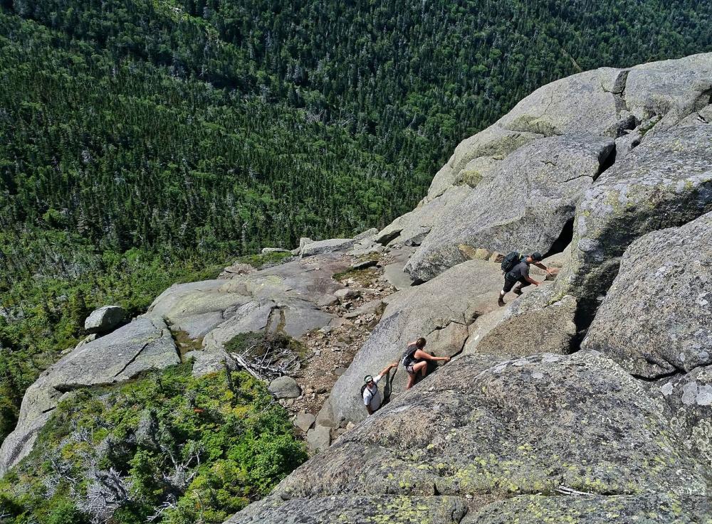 The "Saddleback Cliffs" are a famously challenging section of the Great Range. Three hikers scramble up vertical rocks on the side of a mountain.