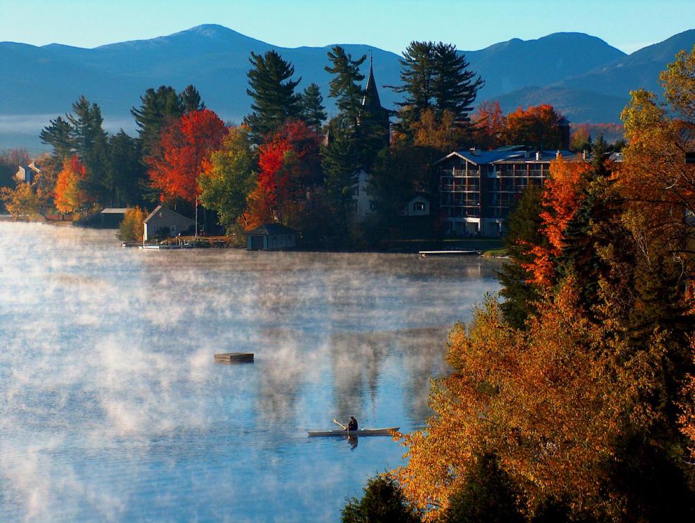 Vibrant autumn colors on the shore of a lake with fog rising over the water and a kayaker paddling.