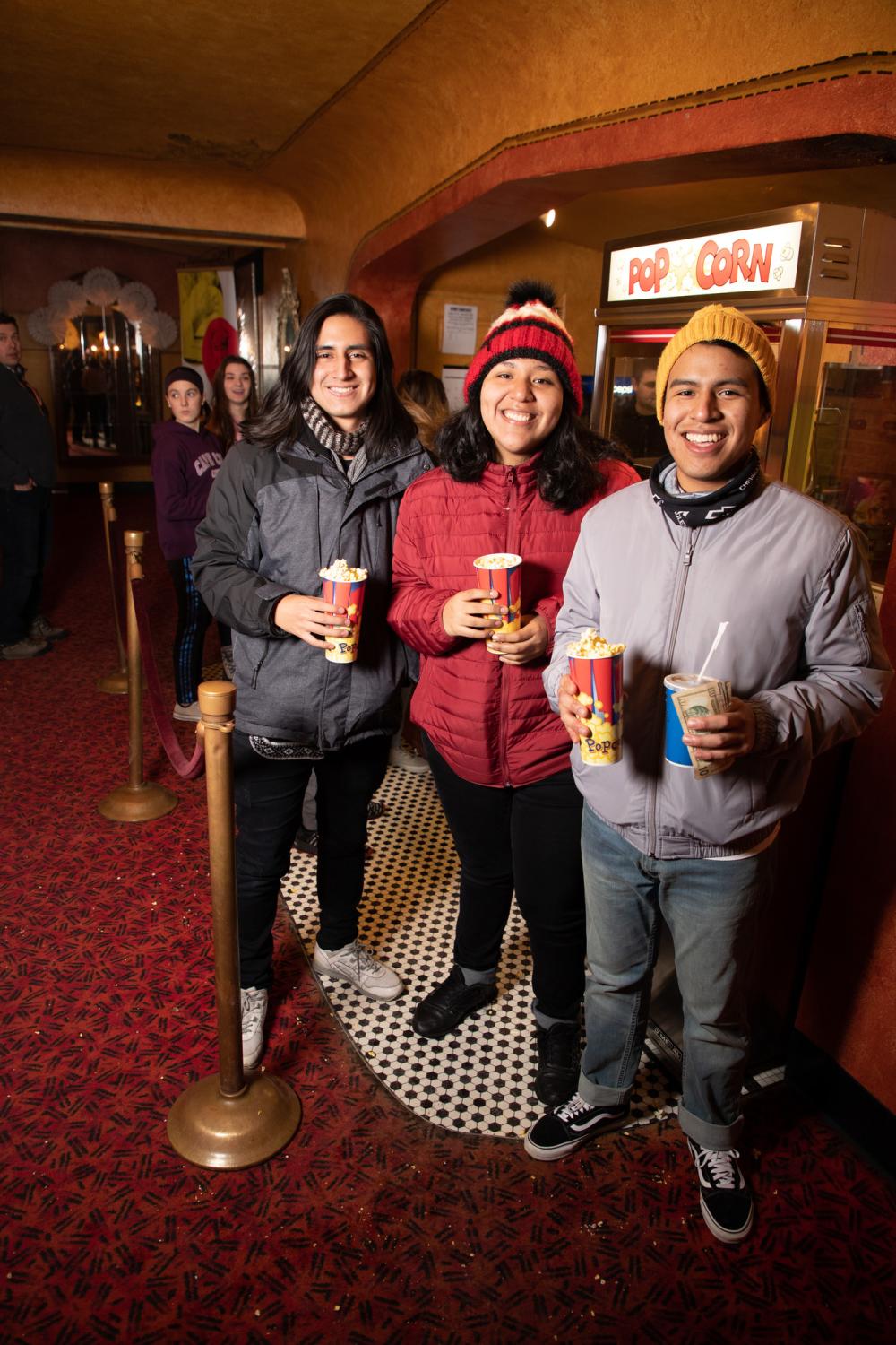 Three people stand in a movie theater lobby, smiling and holding buckets of popcorn.