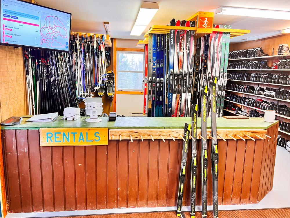 A full rental room with four pairs of skis set out for display. Boots, skis, and poles in the background for rent.