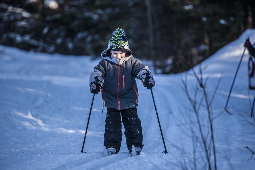 A young boy smiles and cross-country skis on a groomed trail in the woods