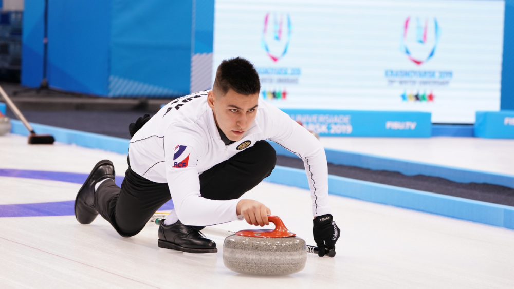 Head to Saranac Lake for the thrills of curling, a winter sports classic! A male curler slides on ice, pushing a curling stone.