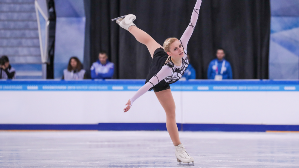 Stunning figure skating competition returns to Lake Placid's Olympic Center! A female figure skater glides across the ice, one leg and arms outstretched.