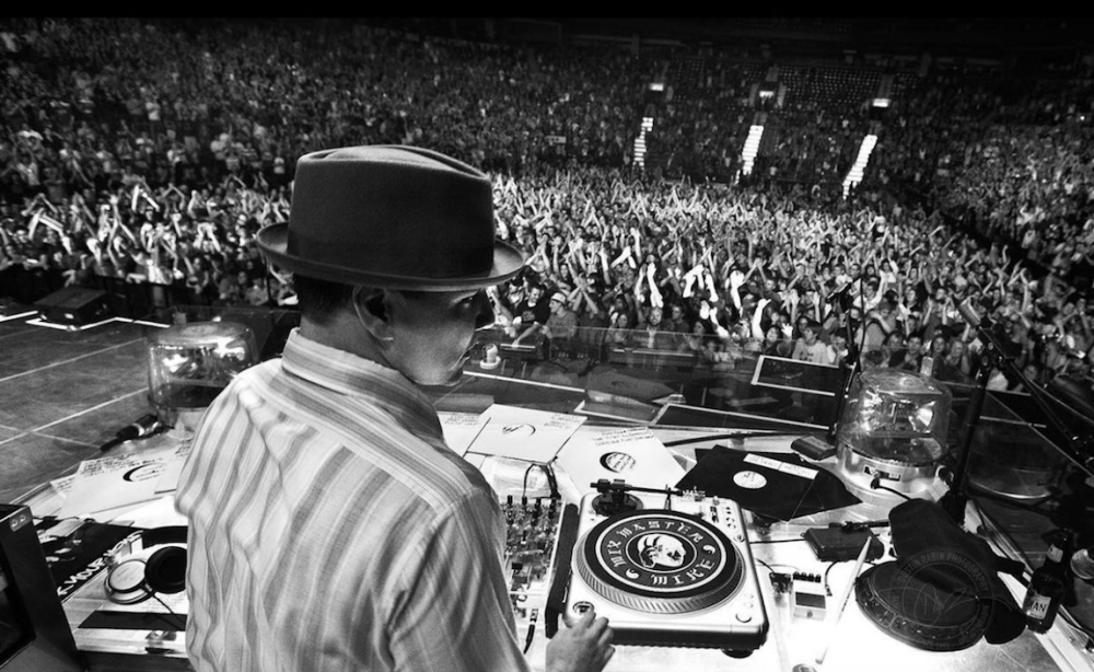 Mix Master Mike will light up Lake Placid! A black and white photo of a DJ wearing a fedora in front of turntables at a concert.