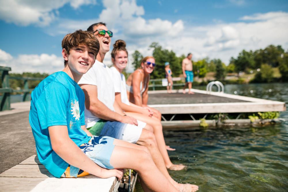 Two adults and a group of teenagers sit in swimming clothing on the edge of a dock in a lake.