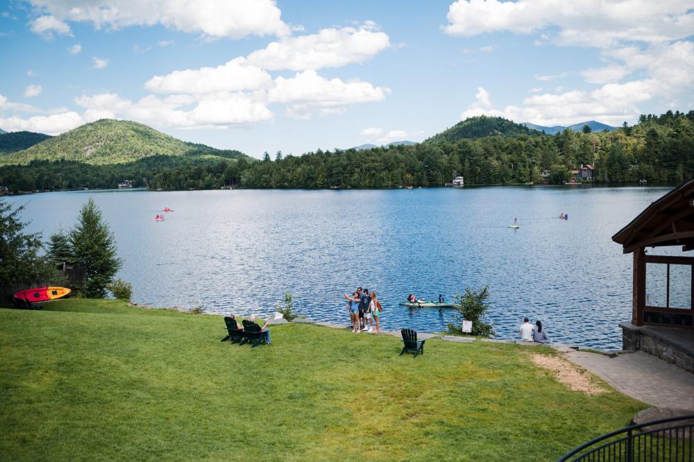 A wide angle of a sloping, grassy park in front of a bright blue lake and mountains. People dot the grass.