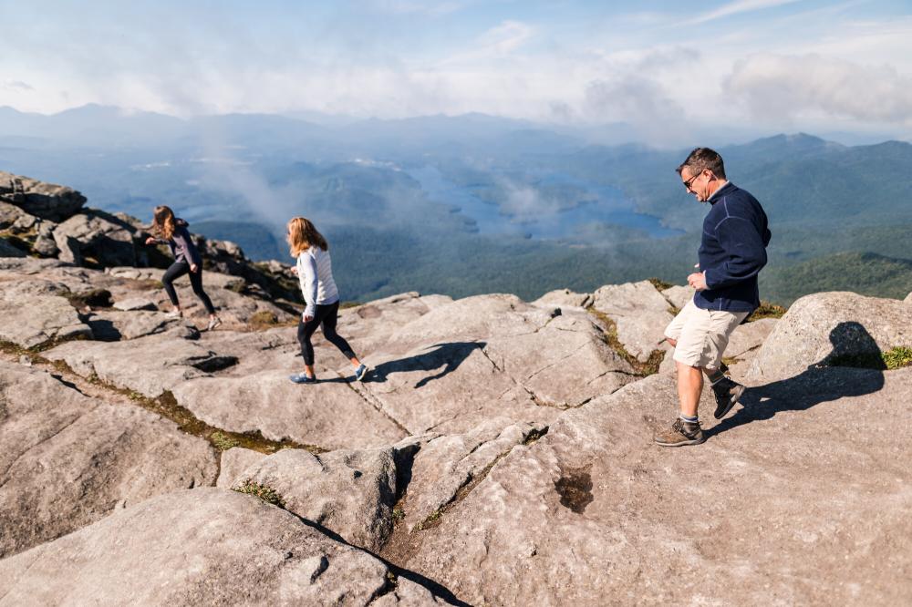 A man and two women in summer clothing walk across a rocky mountain summit on a sunny day.