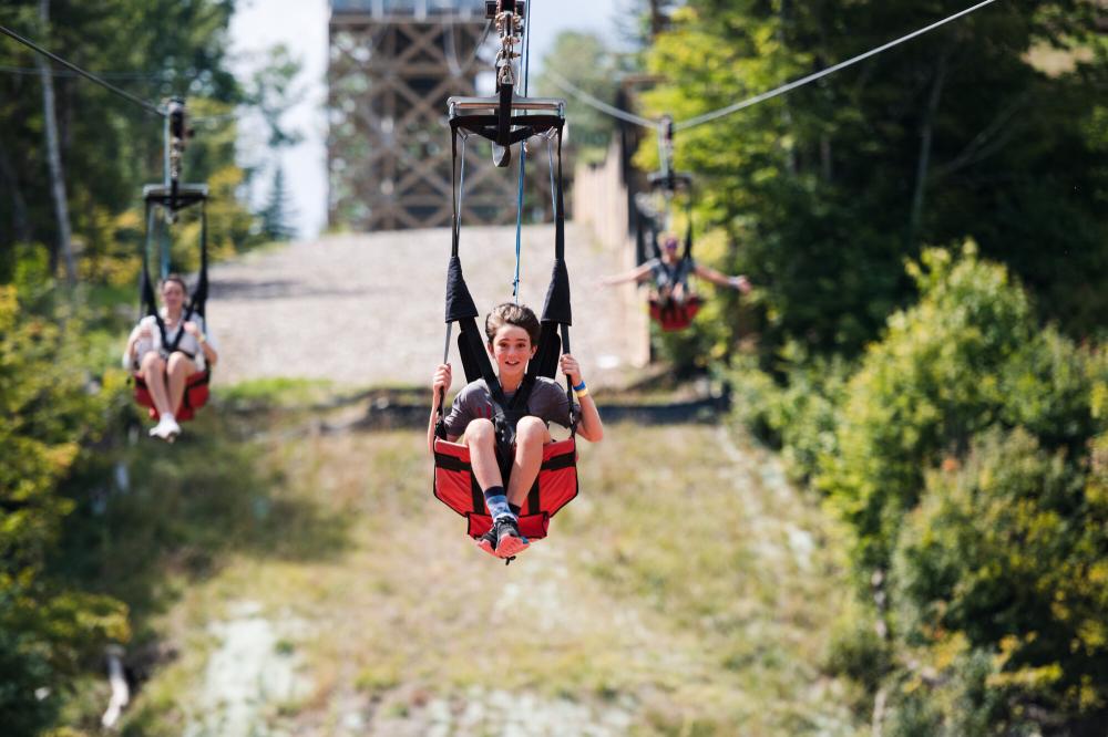 Two teenagers ride down a zipline in red and black harnesses.