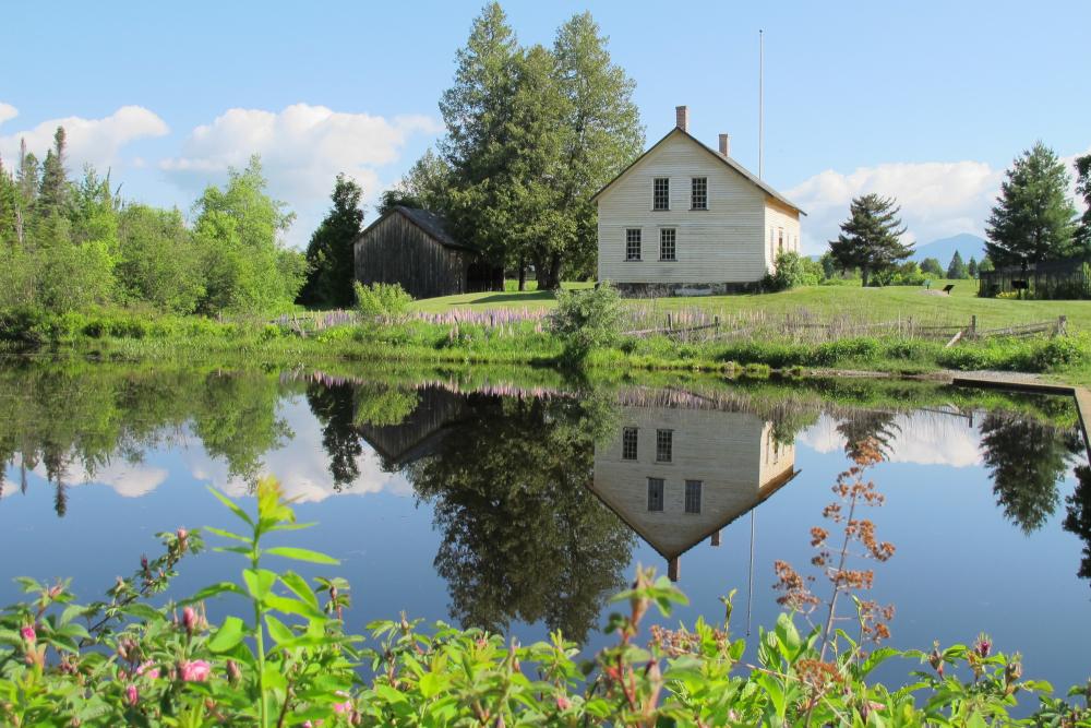 A vintage white farmhouse sits next to a calm pond, surrounded by summer greenery.