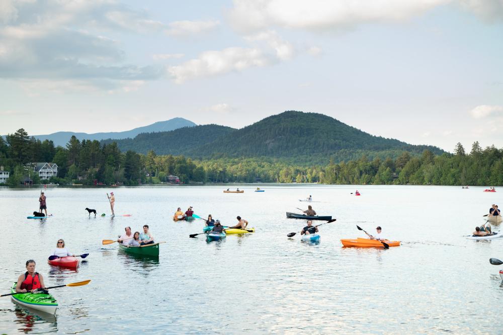 Colorful kayaks, canoes, and SUPs cluster on a sparkling blue lake with green mountains in the background.