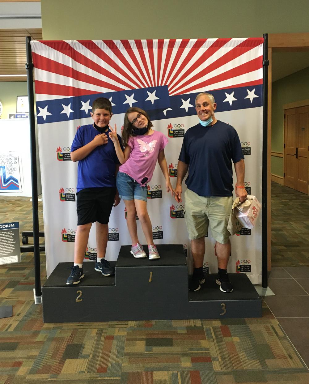 Two children and an adult male stand on a pretend Olympic medal stand in front of a stars and stripes backdrop.