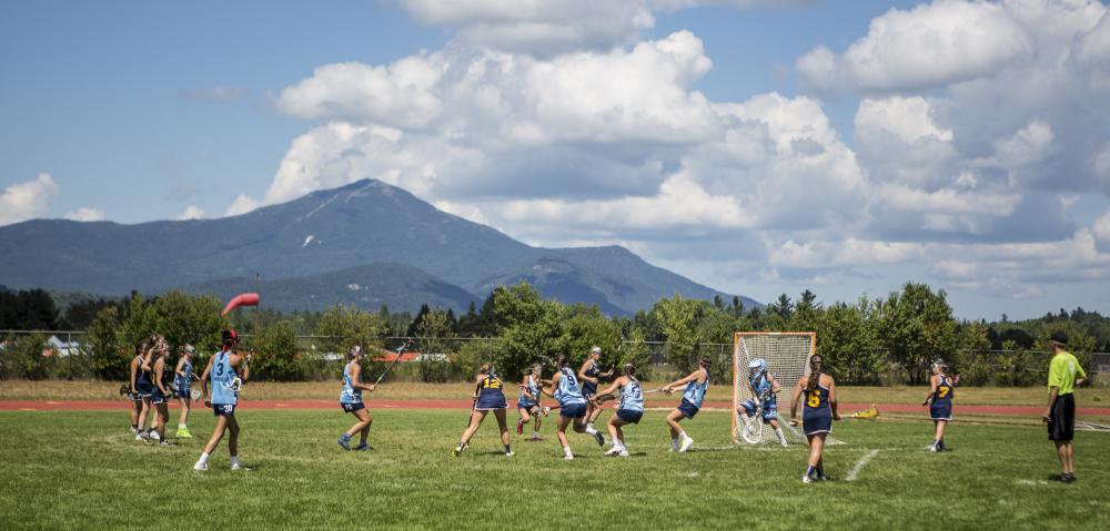 A group of athletes plays lacrosse on an outdoor field in Lake Placid, with high peaks scenery in the distance