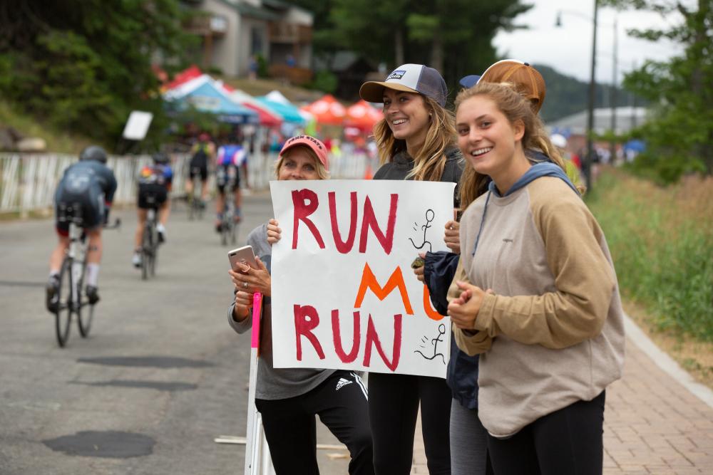Three young women hold a sign with encouraging words for an Ironman athlete at Lake Placid Ironman