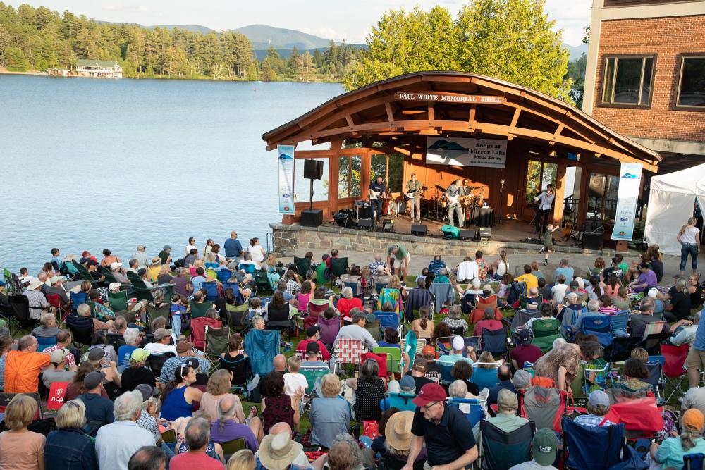 A crowd gathers near the band shell in the grass under a summer sky at Mid's Park in Lake Placid