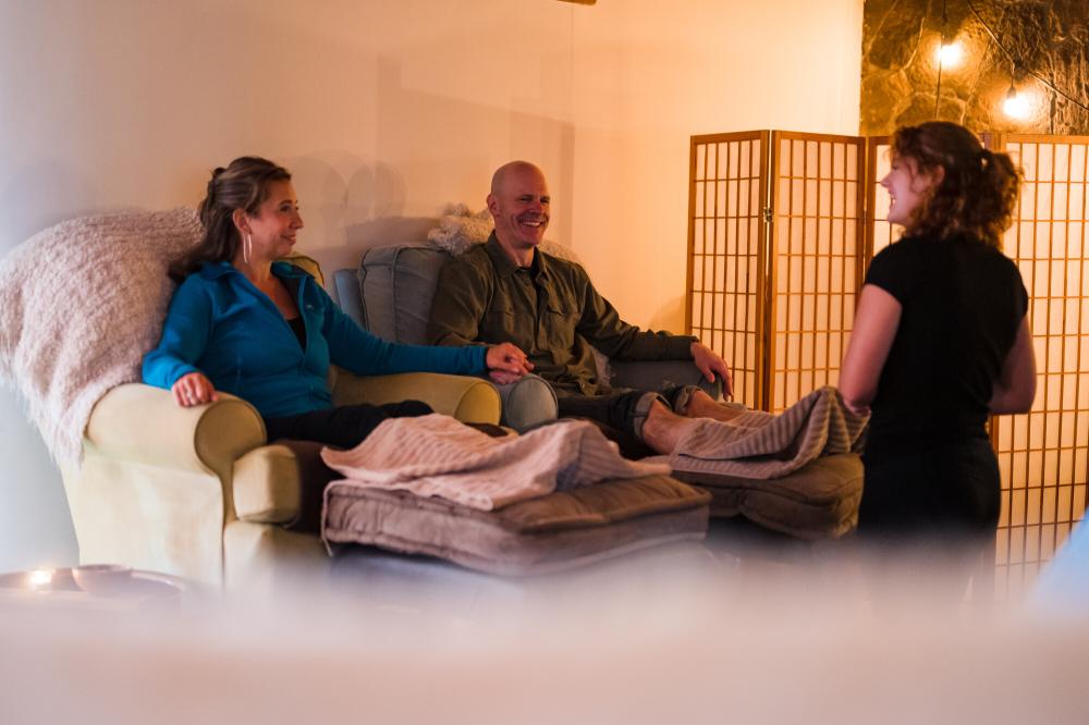 A man and woman sit next to each other on plush armchairs, with their feet up for a spa treatment from a woman with curly hair.