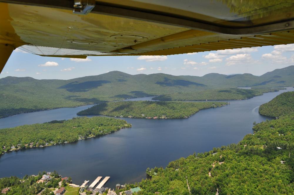 Looking down on green forests, a forested island, and blue lake from under a small yellow airplane wing.