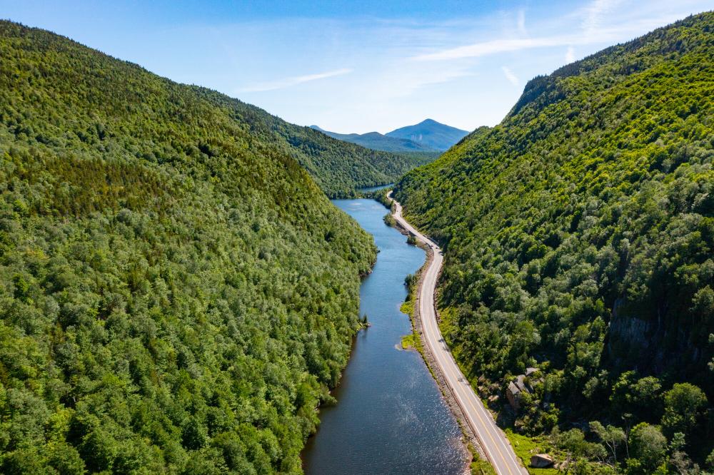 An aerial view of a long, narrow lake, surrounded by a single, narrow road and forested mountains on a sunny day.