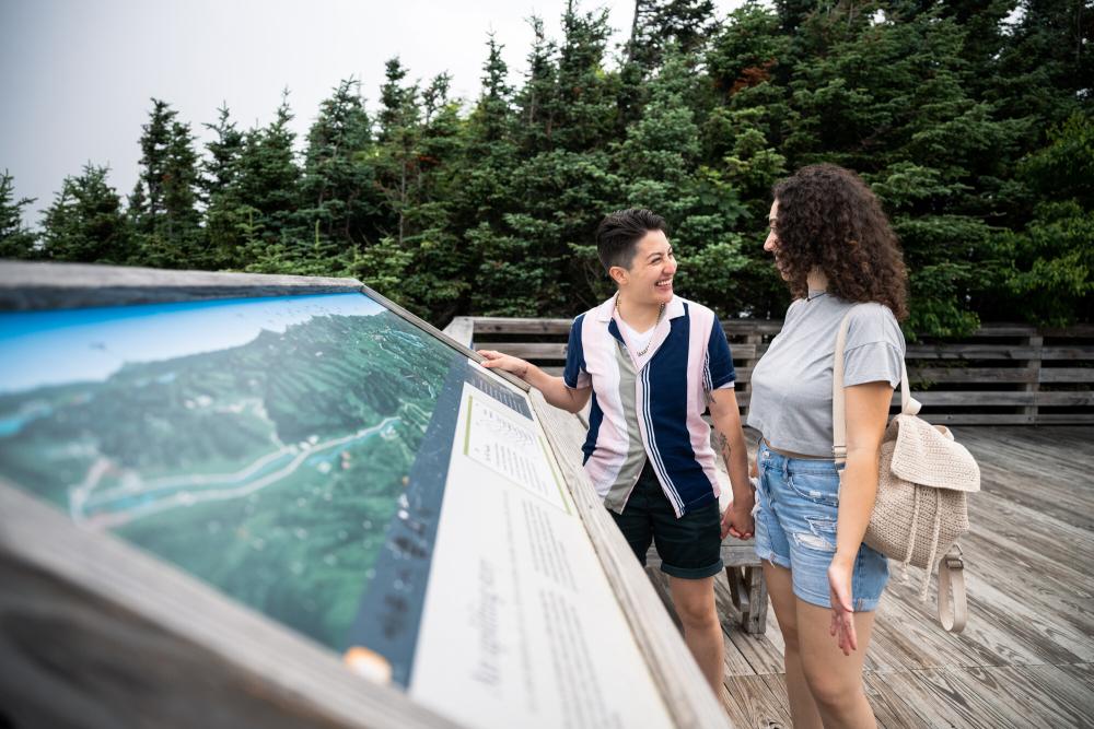 A couple stands smiling at each other in front of an interpretive display on a deck with forest in the background.