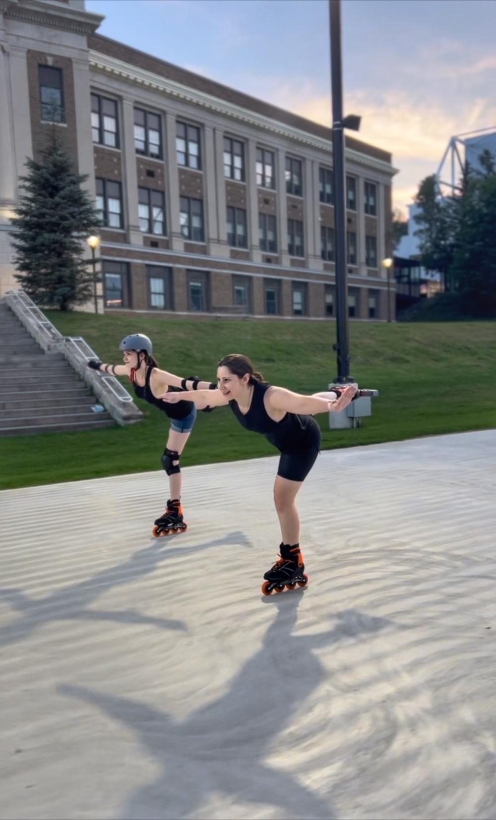 Two women inline skate side-by-side in figure skating poses, skating on one leg.